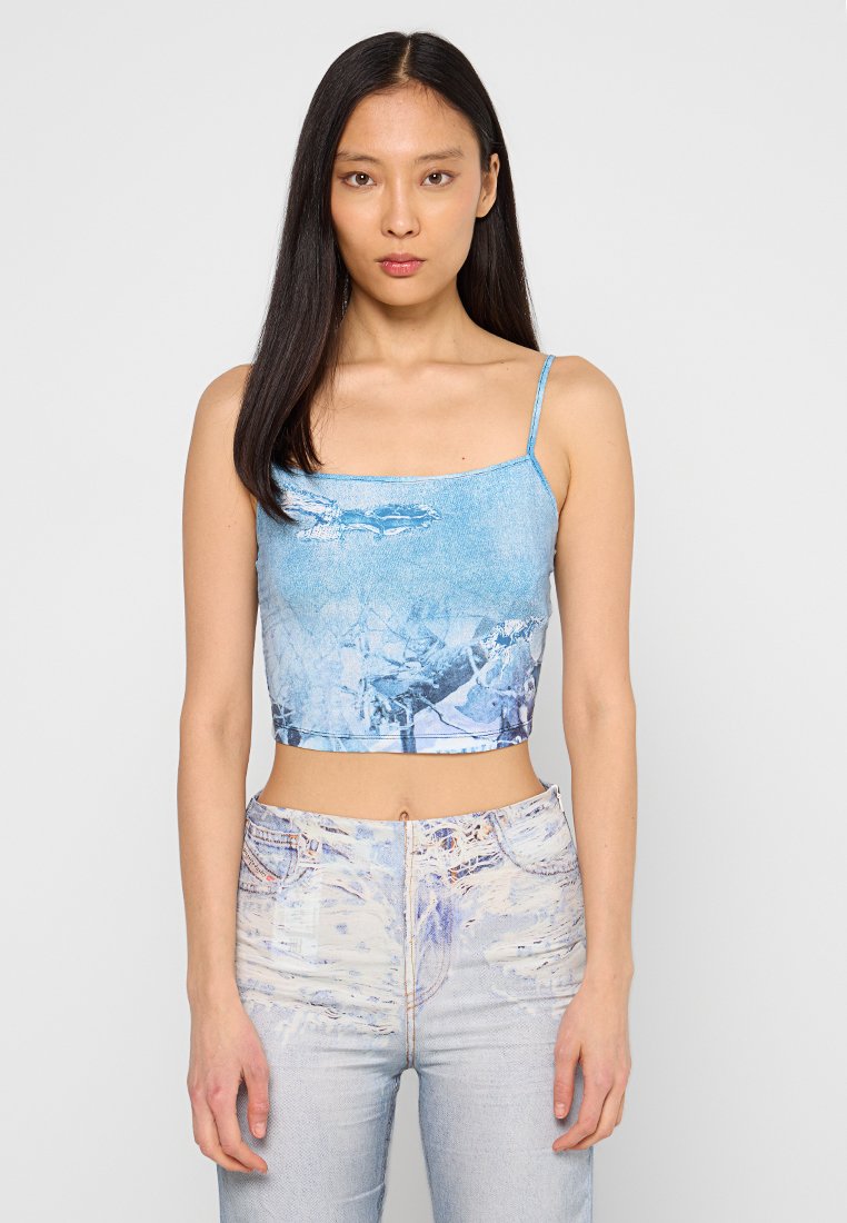 Gekürztes Tanktop aus leicht blauem Stoff mit einem grafischen Druck. Die Träger sind dünn und es hat einen quadratischen Ausschnitt. Texturierte Denim-Jeans runden das Outfit ab.