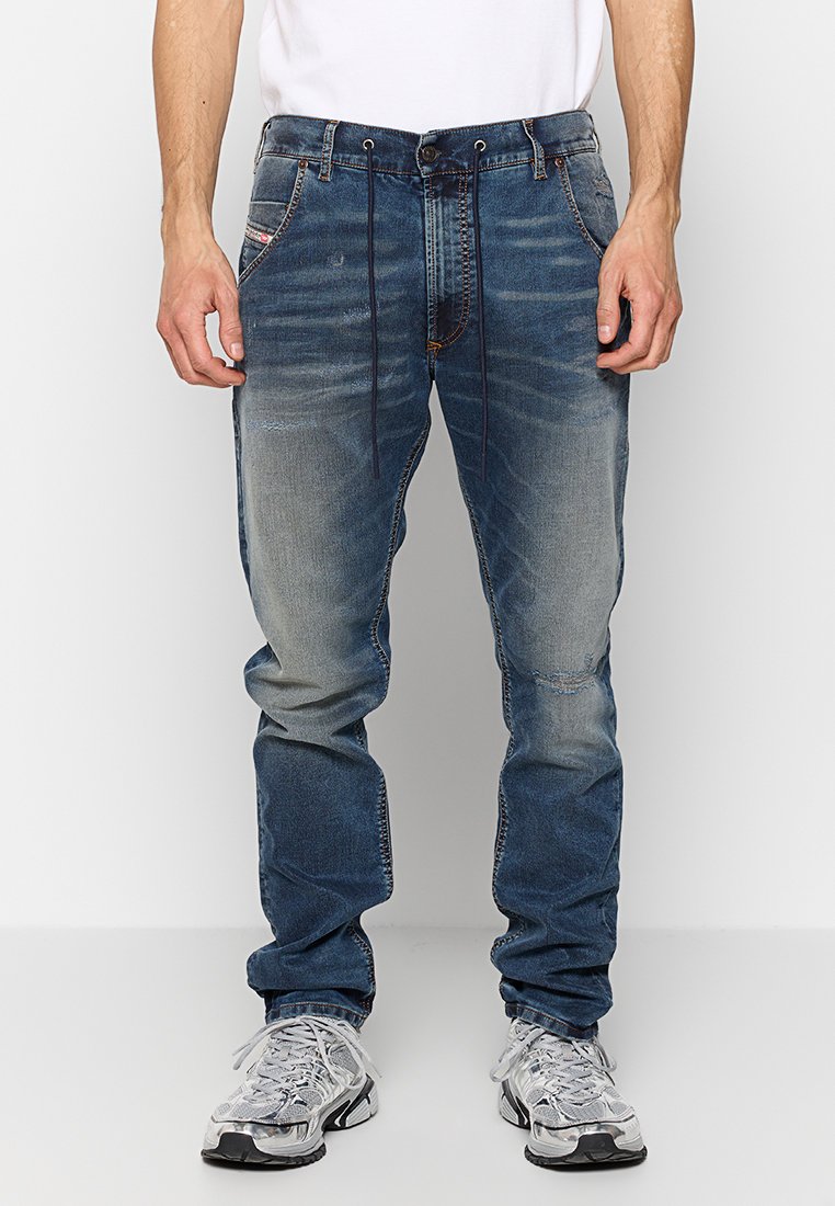Ausgeblichene Jeans in Blau mit geradem Beinschnitt, die subtile Abnutzungsspuren, fünf Taschen und kontrastierende Nähte aufweisen. Getragen mit grauen Sneakers.