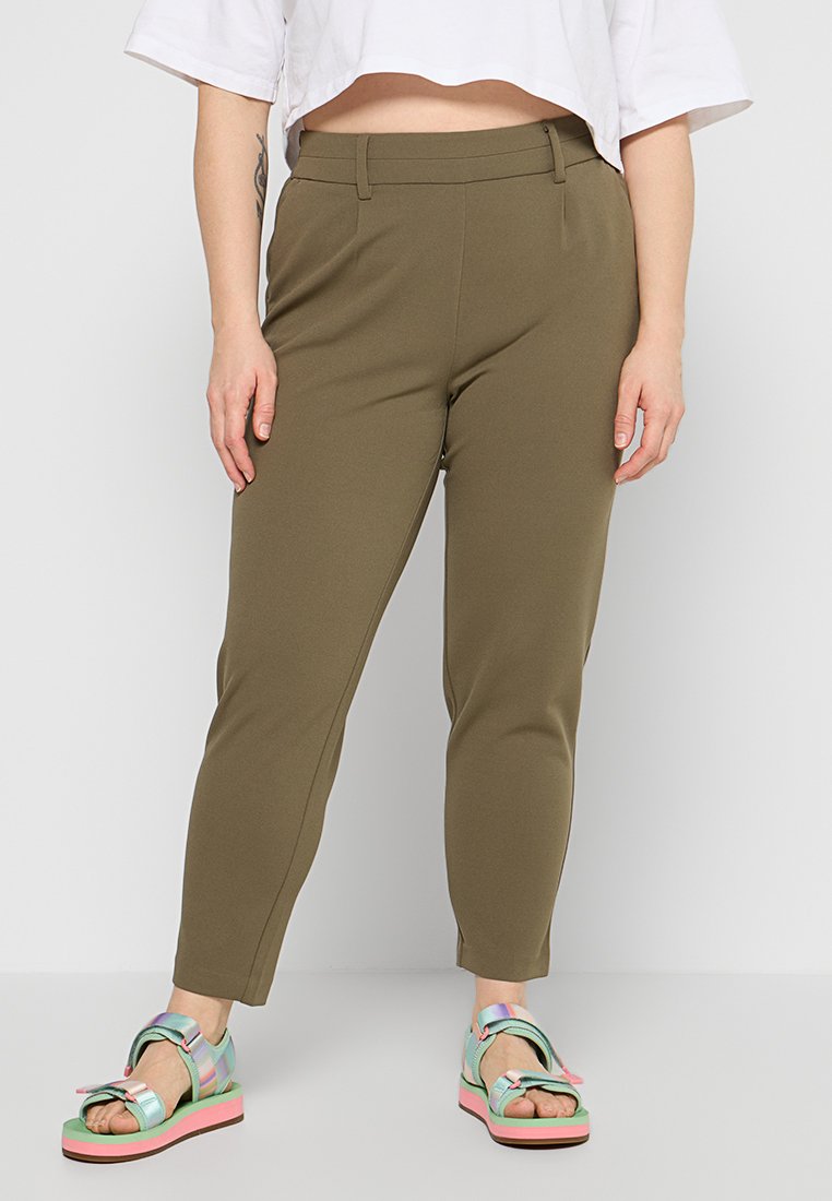 Frau trägt olivgrüne Slim-Fit-Hose und farbenfrohe Plateausandalen, im Stehen vor einem einfarbigen hellen Hintergrund.