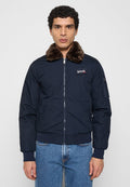 Marineblaue Bomberjacke mit braunem Pelzkragen, Reißverschluss, gerippten Bündchen und Saum sowie seitlichen Taschen. Auf der Brust befindet sich ein Logo-Patch.