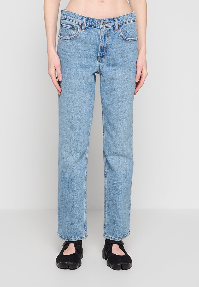 Hellblaue Jeans mit geradem Bein, getragen mit schwarzen Spaltzeh-Schuhen, gezeigt von der Hüfte bis zu den Füßen vor einem hellgrauen Hintergrund.