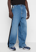 Person in weit geschnittenen blauen Jeans mit schwarzen Seitenstreifen, schwarzem Oberteil und blauen und weißen sportlichen Sneakers, der vor einem weißen Hintergrund steht.