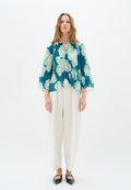Bluse mit floralem Muster in Türkis- und Cremefarben, mit Faltendesign und weiten Ärmeln, kombiniert mit lockeren weißen Hosen und schwarzen Ballerinas.