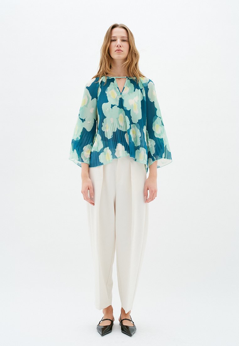 Bluse mit floralem Muster in Türkis- und Cremefarben, mit Faltendesign und weiten Ärmeln, kombiniert mit lockeren weißen Hosen und schwarzen Ballerinas.