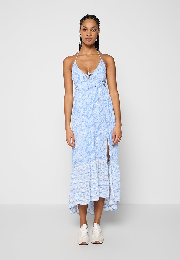 Frau mit Zöpfen, die ein blaues Paisley-Halfter-Kleid mit Frontschlitz und weißen Sneakern trägt, steht vor einem schlichten weißen Hintergrund.