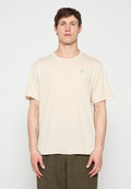 Junger Mann, der ein schlichtes beiges Kurzarm-T-Shirt mit kleinem grünem Logo auf der Brust und strukturierte dunkle Hosen trägt, stehend vor weißem Hintergrund.