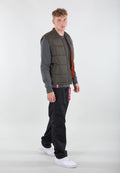Olivgrüner Puffer-Weste mit braunem Innenfutter, Reißverschluss und Seitentaschen, getragen über einem grauen Langarmshirt und schwarzen Hosen. Weiße Sneakers.