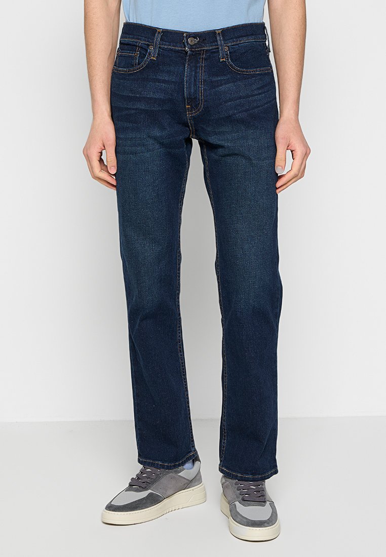 Dunkelblaue Jeans aus Denim mit geradem Schnitt, ausgestattet mit fünf Taschen, einem Knopfverschluss vorne und Kontrastnähten. Dazu werden graue Sneakers getragen.