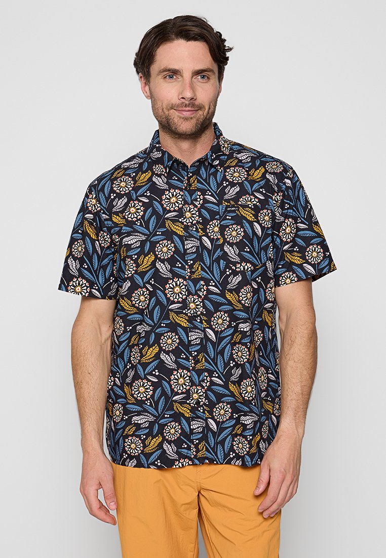 Mann in einem kurzärmligen navyblauen Hemd mit buntem Floralprint und senfgelben Hosen, der vor einem schlichten hellgrauen Hintergrund steht.