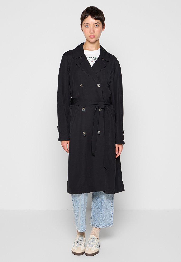Schwarzer Trenchcoat mit doppelreihigen Knöpfen, tailliertem Gürtel und langen Ärmeln. Getragen über einem weißen Hemd und kombiniert mit hellen Jeans.