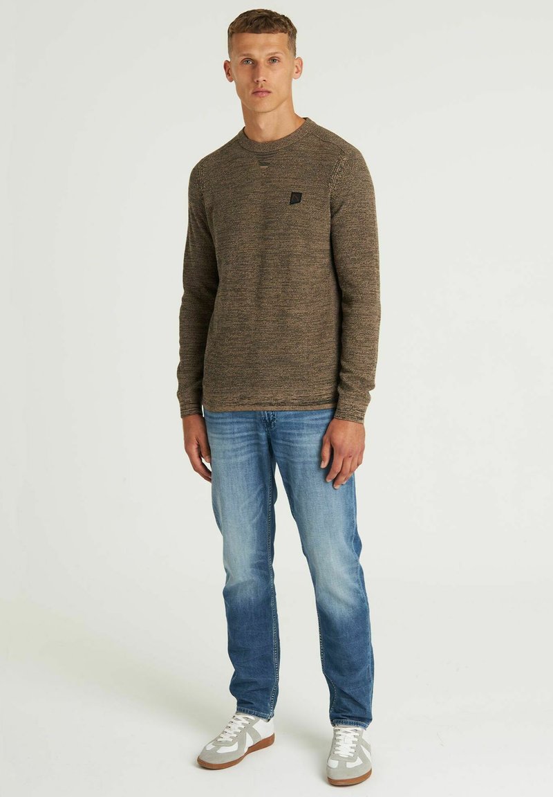 Brauner Strickpullover mit rundem Ausschnitt, einem kleinen schwarzen Logo-Patch, kombiniert mit hellblauen Jeans und grauen Sneakers mit weißen Akzenten.