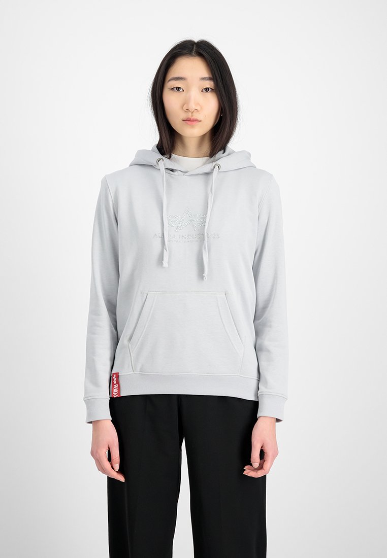 Junge Frau steht mit Blick nach vorne, trägt einen hellgrauen Hoodie mit Kängurutasche und schwarze Hose vor einem neutralen Hintergrund.