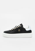 Schwarzer Sneaker mit niedrigem Schaft, weißen Schnürsenkeln, goldenen Ösen, weißem Fersenlaschen, goldenem Logo an der Seite und dicker weißer Plateausohle.