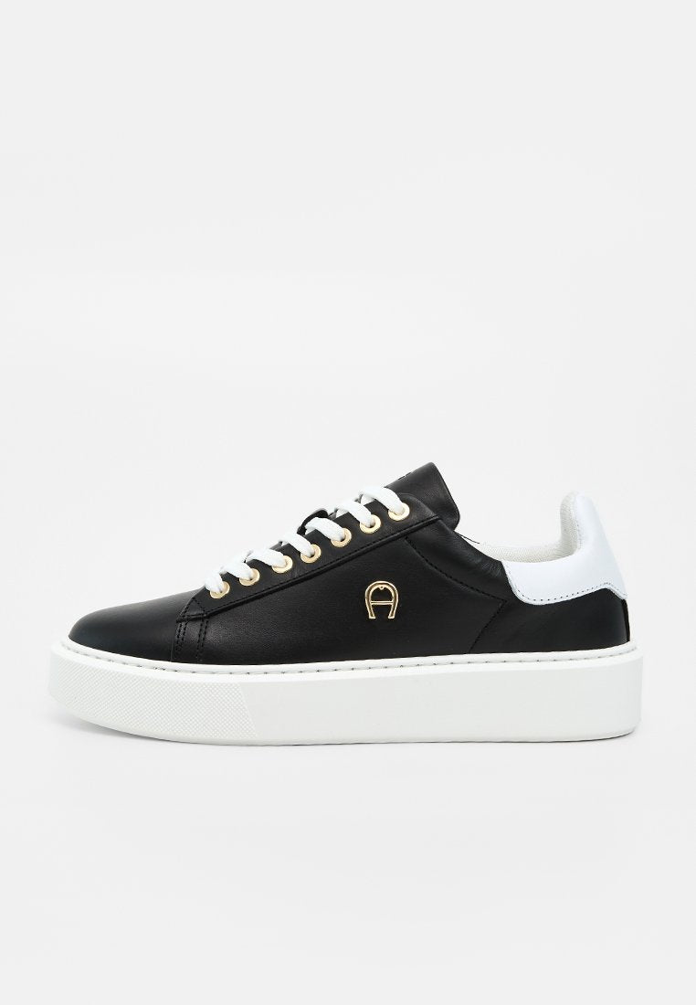 Schwarzer Sneaker mit niedrigem Schaft, weißen Schnürsenkeln, goldenen Ösen, weißem Fersenlaschen, goldenem Logo an der Seite und dicker weißer Plateausohle.