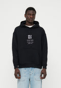 Mann trägt schwarzen Hoodie mit weißem "HUGO"-Schriftzug und hellblauen Jeans, steht vor einem schlichten weißen Hintergrund.
