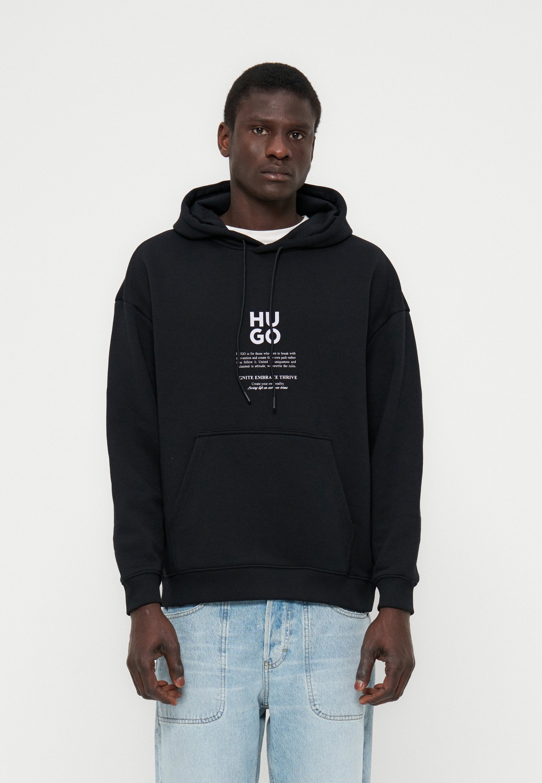 Mann trägt schwarzen Hoodie mit weißem "HUGO"-Schriftzug und hellblauen Jeans, steht vor einem schlichten weißen Hintergrund.