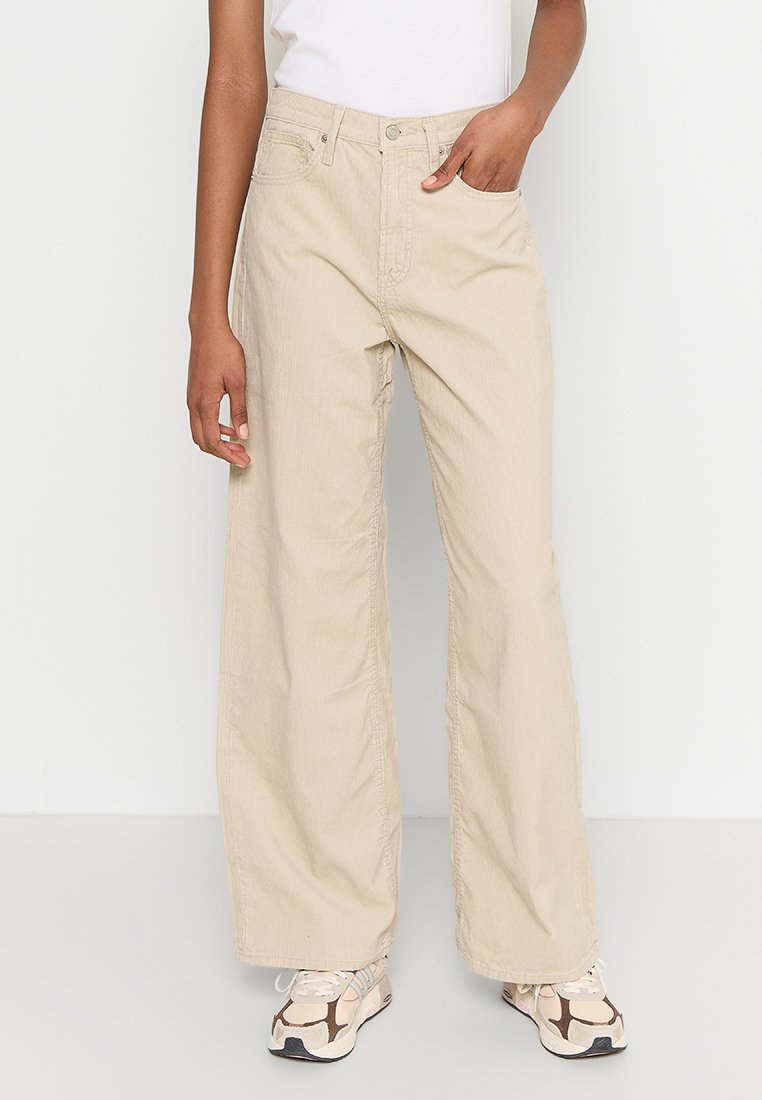 Person in beigen, weiten Hosen, weißen Sneakers und einem in die Hose gesteckten weißen ärmellosen Oberteil, der vor einem schlichten weißen Hintergrund steht.