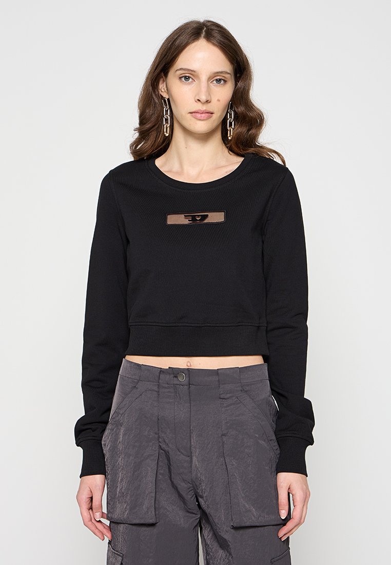 Schwarzer, kurzer Sweatshirt mit gerippter Textur, ausgestattet mit einem rechteckigen braunen Logo-Patch in der Mitte. Lange Ärmel und ein runder Halsausschnitt.