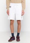 Mann trägt weiße Shorts mit Kordelzug und Patches an den Seiten, ein beige-farbenes Langarmoberteil, dunkelblaue Socken und kastanienbraune Sneakers mit blauen Streifen.