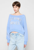 Frau mit kurzen braunen Haaren, die ein hellblaues schulterfreies Sweatshirt mit der Aufschrift "MAKING WAVES" und hellblaue Jeans trägt.