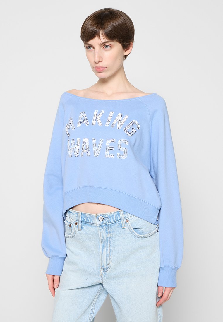 Frau mit kurzen braunen Haaren, die ein hellblaues schulterfreies Sweatshirt mit der Aufschrift "MAKING WAVES" und hellblaue Jeans trägt.