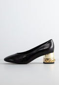Schwarzer Lackleder-Niedrigabsatzschuh mit goldfarbener, metallic-blokkierten Absatz, die mit runden Nieten verziert ist, auf einer schlichten weißen Oberfläche präsentiert.