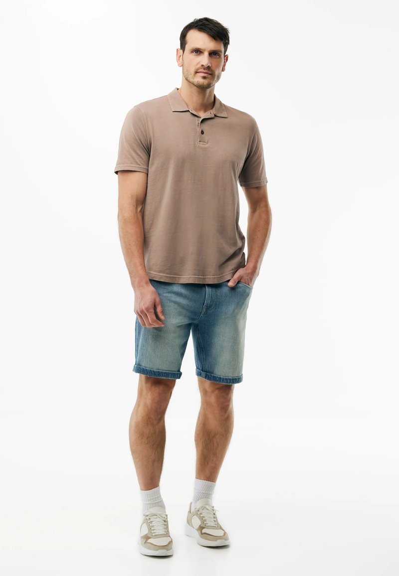 Mann steht mit einer Hand in der Tasche, trägt ein braunes Poloshirt, Jeansshorts, weiße Socken und beige Turnschuhe vor weißem Hintergrund.
