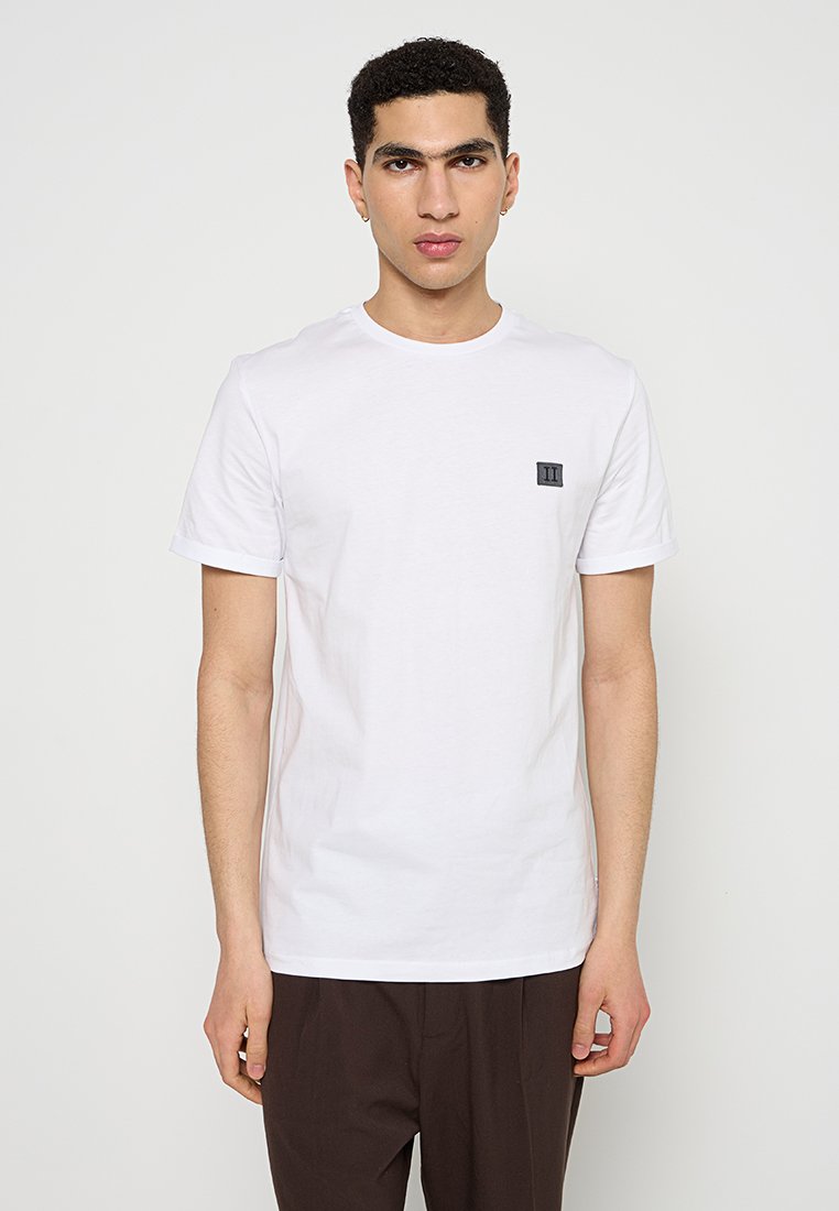 Weißes T-Shirt aus Baumwolle mit kurzen Ärmeln, Rundhalsausschnitt und einem kleinen schwarzen Logopatch auf der linken Brust. Glatte Textur und normale Passform.
