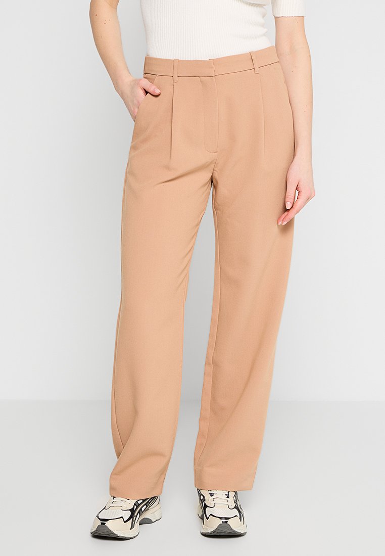 Hellbraune maßgeschneiderte Hose mit hoher Taille, Falten und geradem Schnitt, kombiniert mit einem weißen Strickoberteil und schwarz-weißen Sneakern.