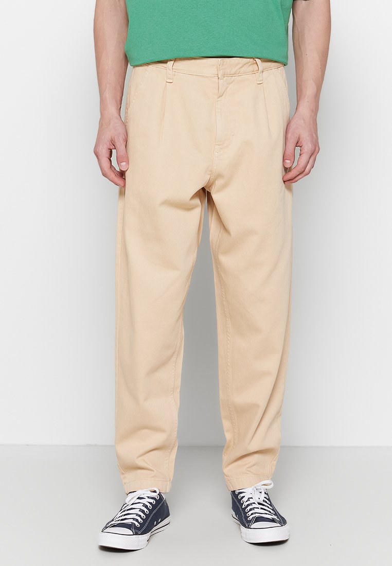 Beige Chinos mit einem lockeren Schnitt, ausgestattet mit einer flachen Vorderseite, Gürtelschlaufen und Seitentaschen, kombiniert mit blauen Sneakers.