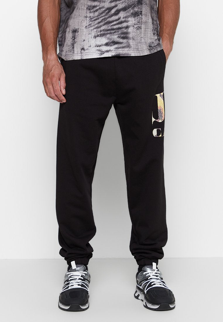 Mann in schwarzen Jogginghosen mit buntem Logo auf dem Oberschenkel und schwarz-weißen und grauen Laufschuhen, der vor weißem Hintergrund steht.