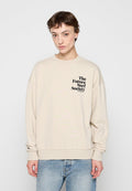 Person mit kurzen braunen Haaren, die einen beigen Sweatshirt mit dem Schriftzug „The Future Surf Society“ und hellblaue Jeans vor einem einfarbigen Hintergrund trägt.