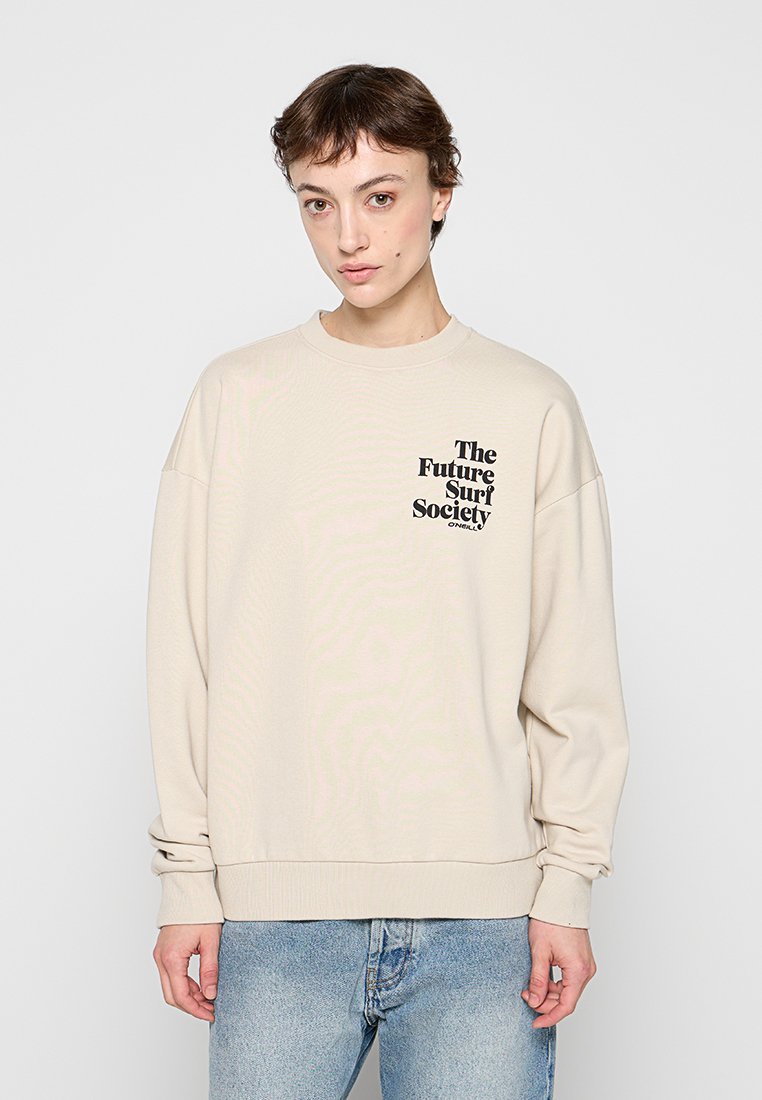 Person mit kurzen braunen Haaren, die einen beigen Sweatshirt mit dem Schriftzug „The Future Surf Society“ und hellblaue Jeans vor einem einfarbigen Hintergrund trägt.