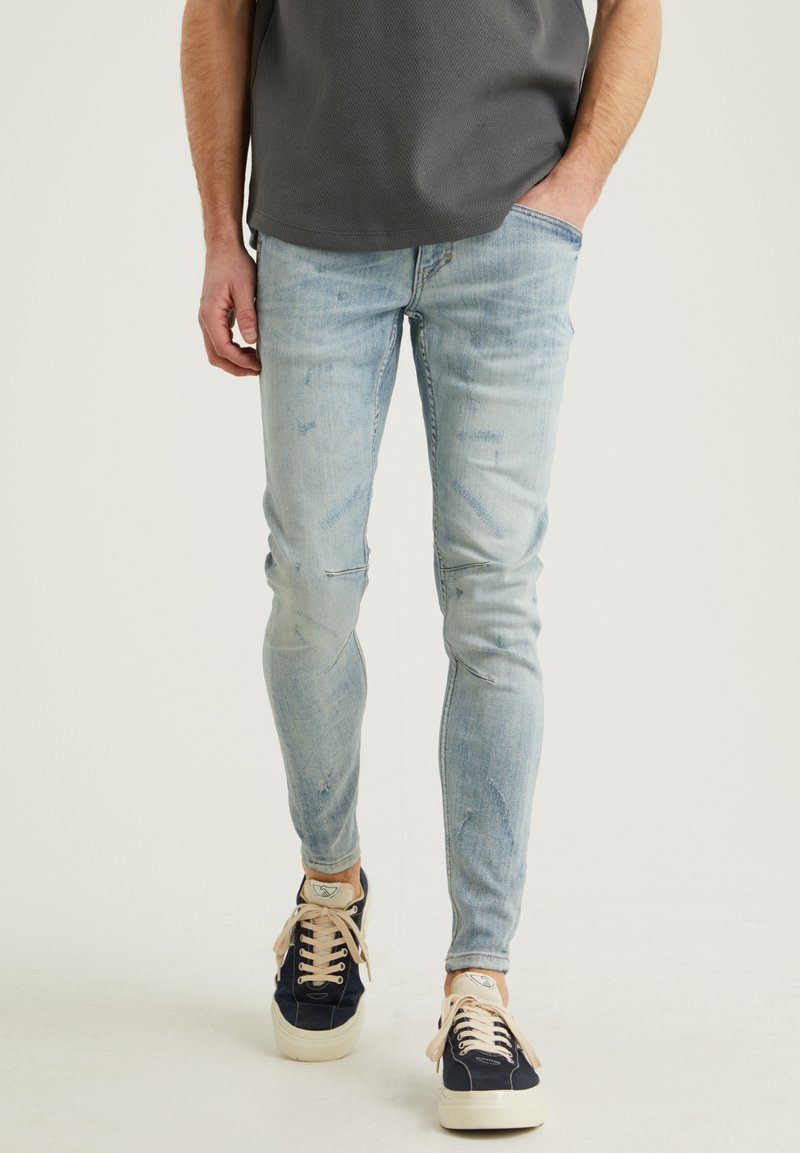 Hellblaue Slim-Fit-Jeans mit Verwaschungen und Used-Look-Details, kombiniert mit marineblauen Sneakers mit beigen Schnürsenkeln und einer strukturierten Oberfläche.