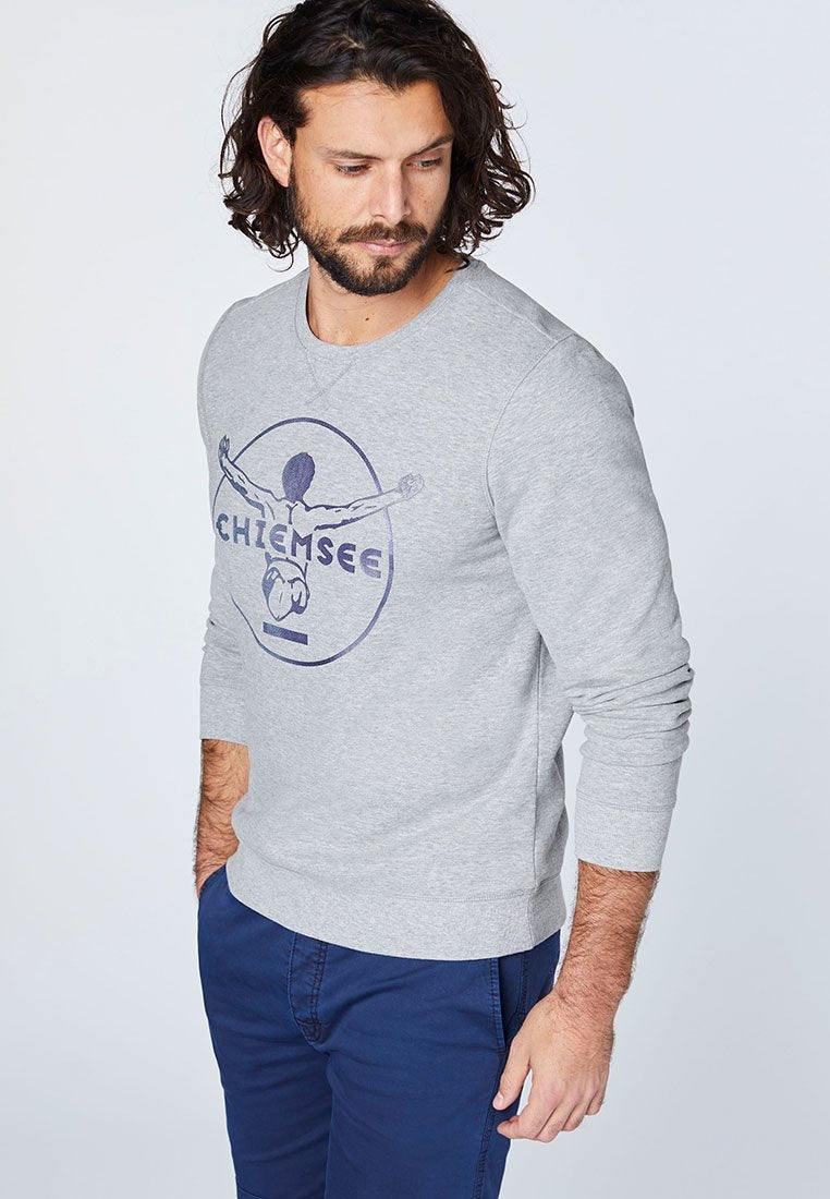 Grauer Sweatshirt mit langen Ärmeln, das einen grafischen Druck in Dunkelblau aufweist. Gerippter Halsausschnitt, Bündchen und Saum. Kombiniert mit blauen Hosen.