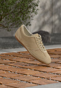 Beiger Wildledersneaker mit Schnürsenkeln, der über einem Holzkachelboden schwebt, mit Grünpflanzen und einer grauen Wand im Hintergrund.