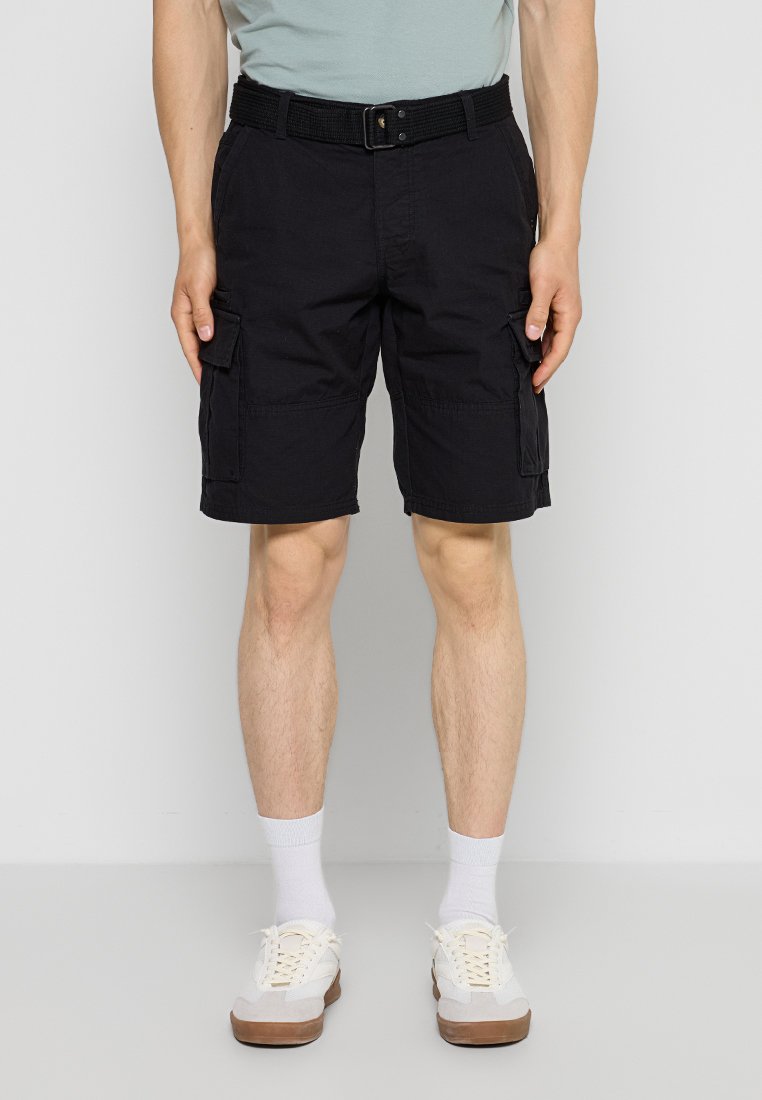 Mann trägt schwarze Cargo-Shorts mit Seitentaschen, einen schwarzen Gürtel, weiße Socken und weiße Turnschuhe, steht vor einem schlichten hellen Hintergrund.