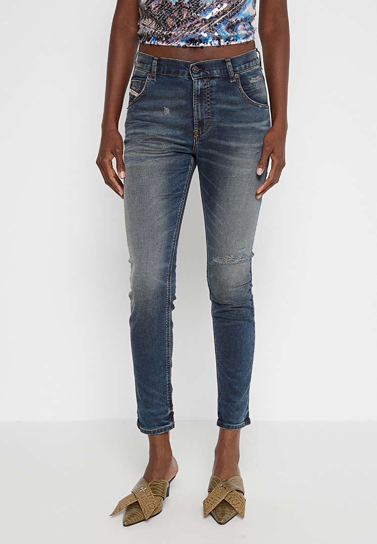 Ausgeblichene blaue Denim-Jeans mit schmaler Passform, die über Distressed-Details und ein traditionelles Five-Pocket-Design verfügen. Getragen mit grünen, gemusterten Slippern.