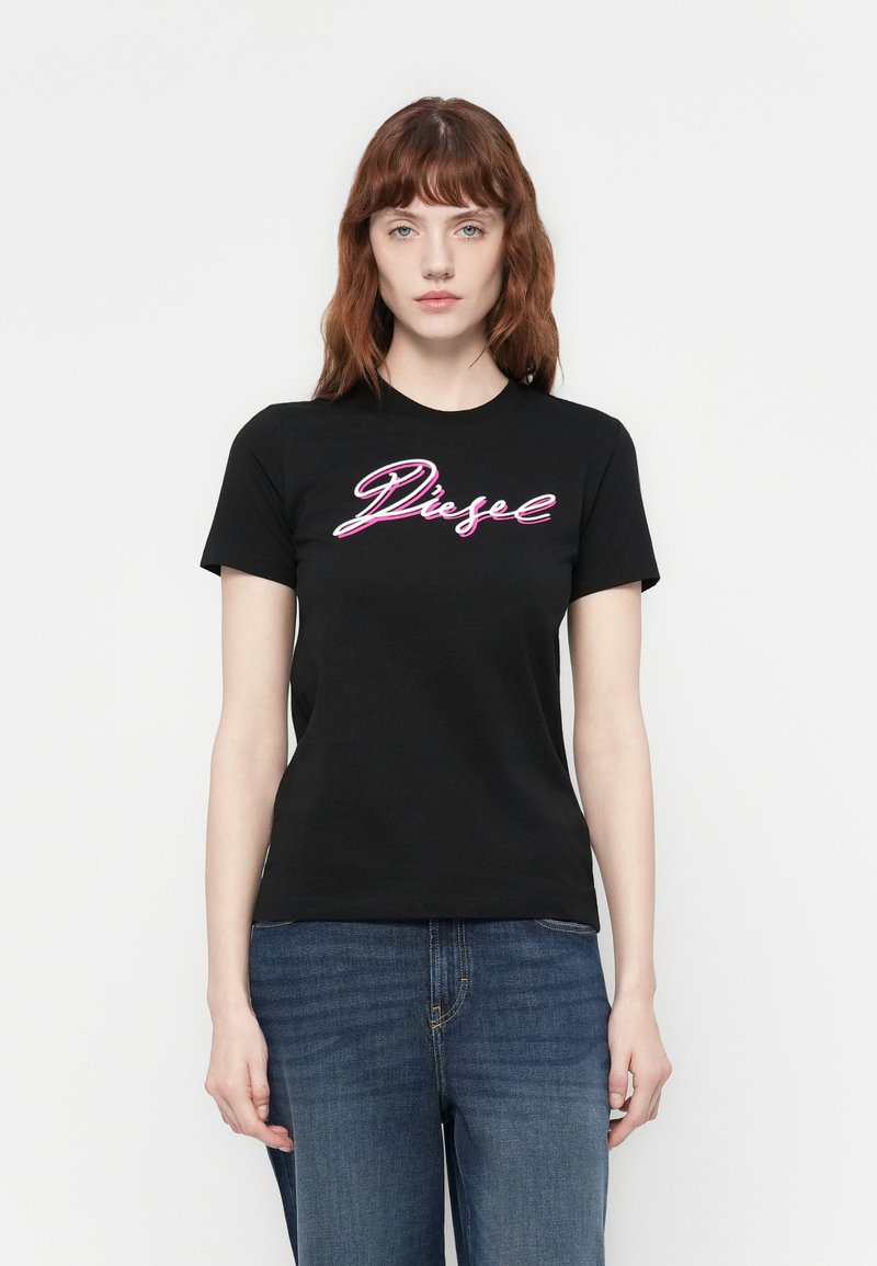 Junge Frau mit roten Haaren trägt ein schwarzes Diesel-T-Shirt und blaue Jeans und steht vor einem schlichten weißen Hintergrund.