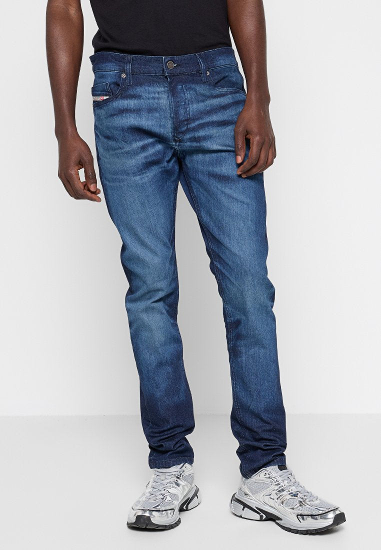Mann in eng anliegenden blauen Jeans und silbernen Sportschuhen, der vor einem schlichten weißen Hintergrund steht.