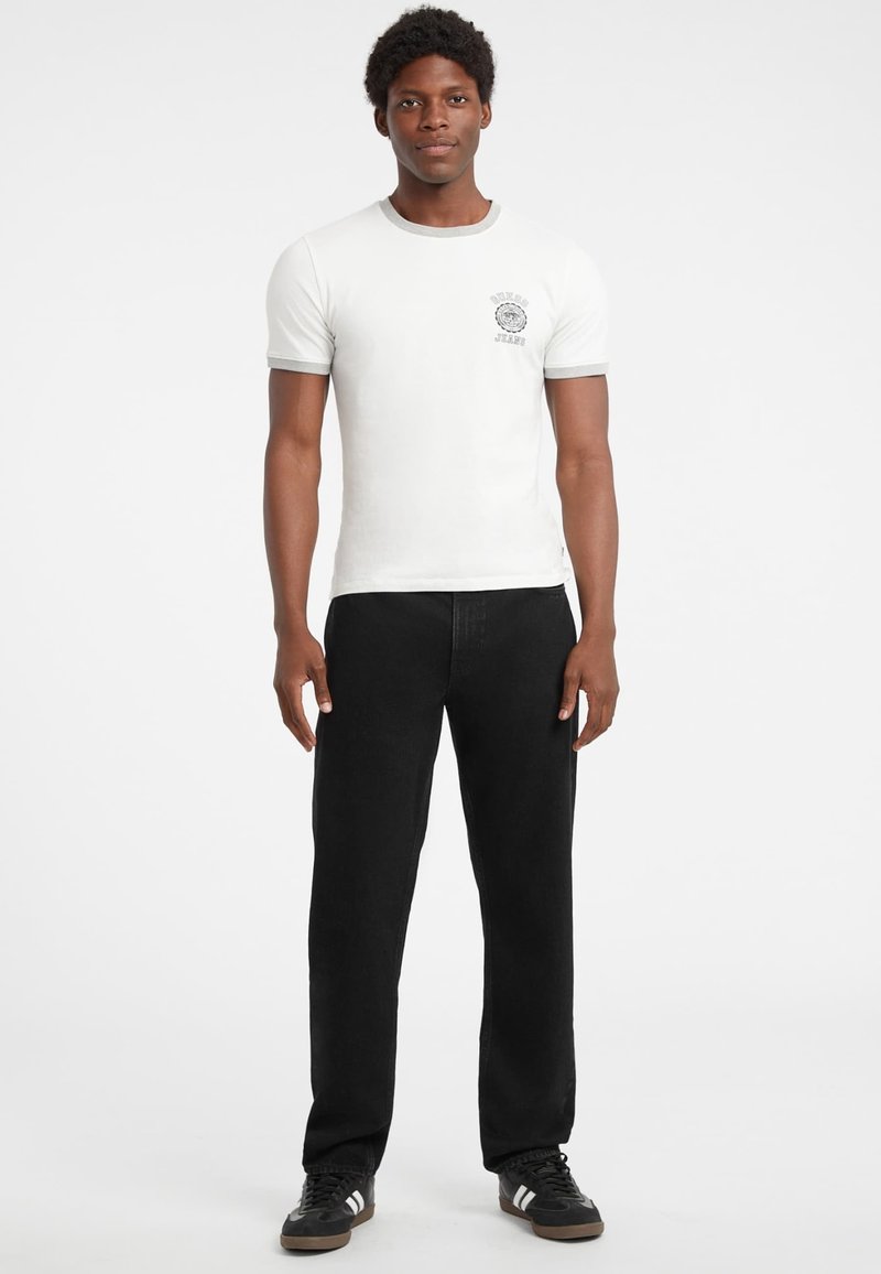 Weißes Baumwoll-T-Shirt mit grauen Akzenten und Logo auf der Brust, kombiniert mit schwarzen Denim-Hosen und schwarzen Sneakers. Gerade geschnittener Beinverlauf.