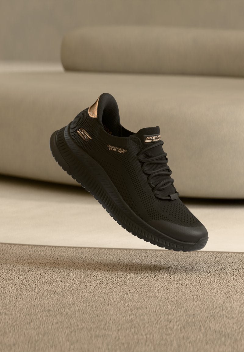Schwarzer Skechers Slip-On-Sneaker mit Mesh-Obermaterial und roségoldfarbenen Akzenten, schwebt über einem beigen Teppich vor einem beigen Sofa.