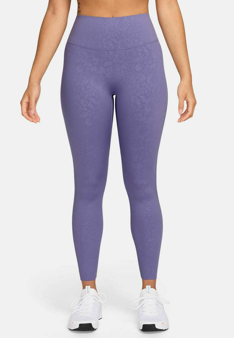 Lila Leggings mit einem strukturierten floralen Muster. Hoher Bund; glatter, dehnbarer Stoff. Getragen mit weißen Sportschuhen.