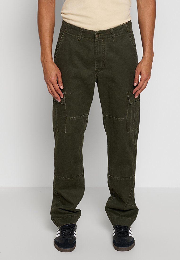 Cargo-Hosen aus dunkelgrünem Baumwollstoff. Mit seitlichen Taschen und einer entspannten Passform, sichtbaren Nähten und einer Knopfverschlüsse. Schwarze Sneakers darunter.