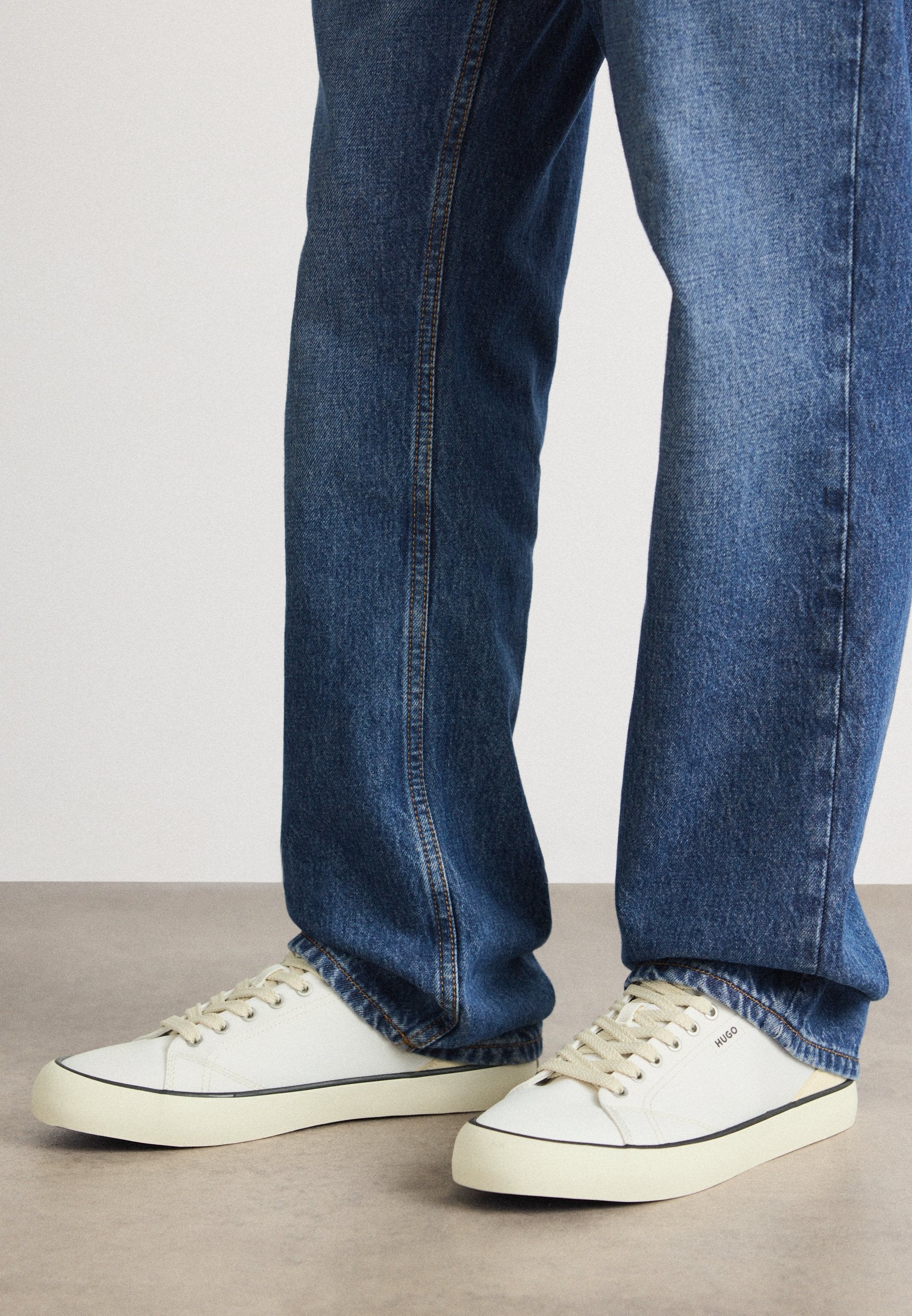 Weiße Canvas-Sneaker mit beigen Schnürsenkeln und schwarzen Akzenten, kombiniert mit blauen Jeans im geraden Schnitt und heller Waschung.