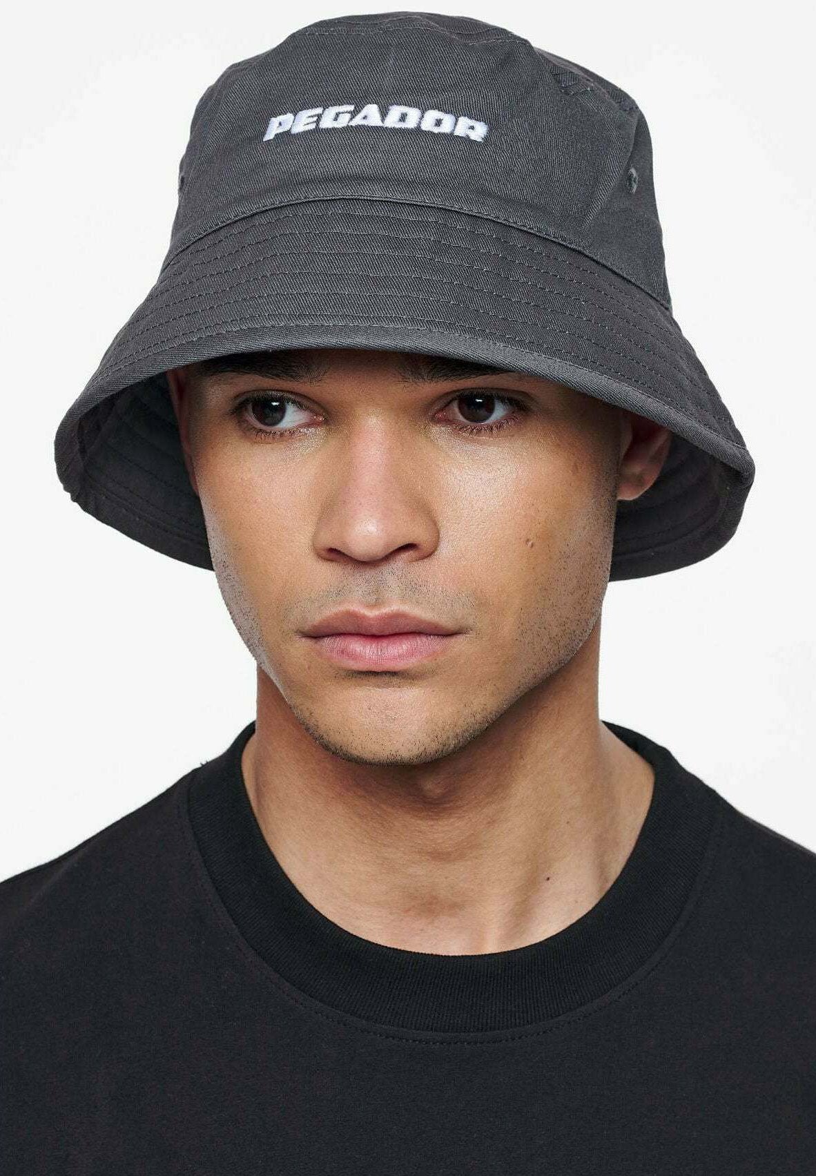 Grau Bucket Hat mit "PEGADOR"-Logo in Weiß, aus weichem Stoffmaterial und breitem, nach unten geneigtem Rand. Getragen von einem Modell mit einem schwarzen Shirt.