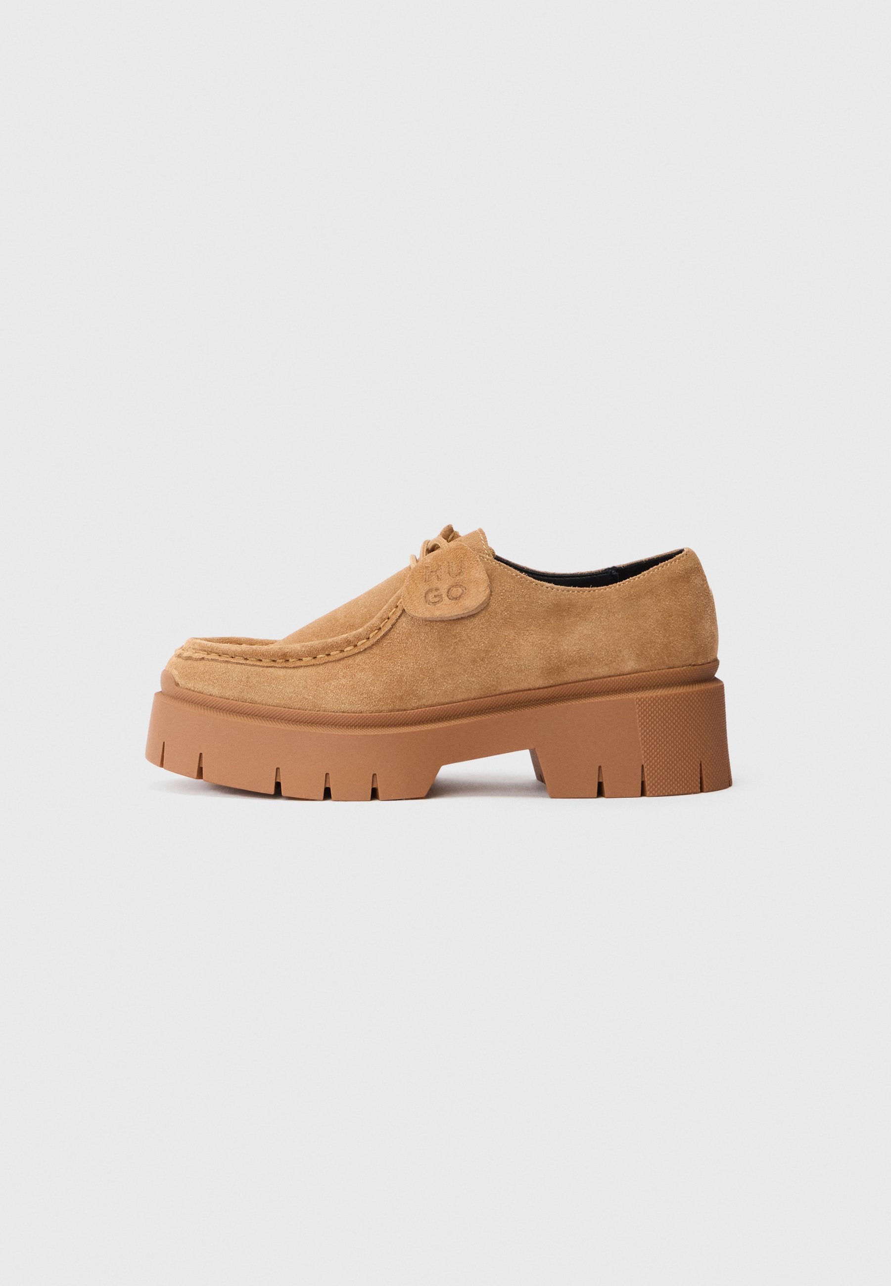 Beige Wildleder-Plattform-Pantolette mit dicker Sohle und gestickten Details, ausgestattet mit einem kleinen Etikett, das nahe den Schnürsenkeln mit "HUGO" geprägt ist.