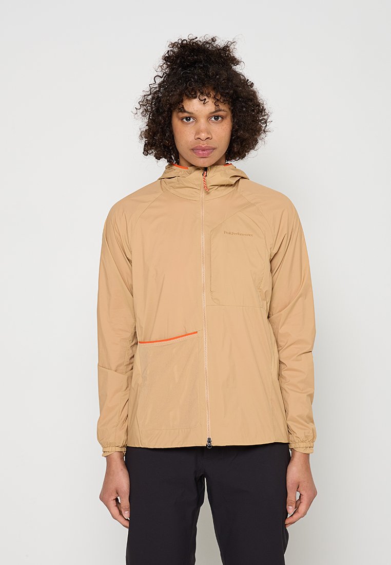 Person mit lockigem Haar, die eine beige Kapuzenjacke mit Frontreißverschluss und orange umrandeter Tasche trägt, steht vor einem einfarbigen hellen Hintergrund.