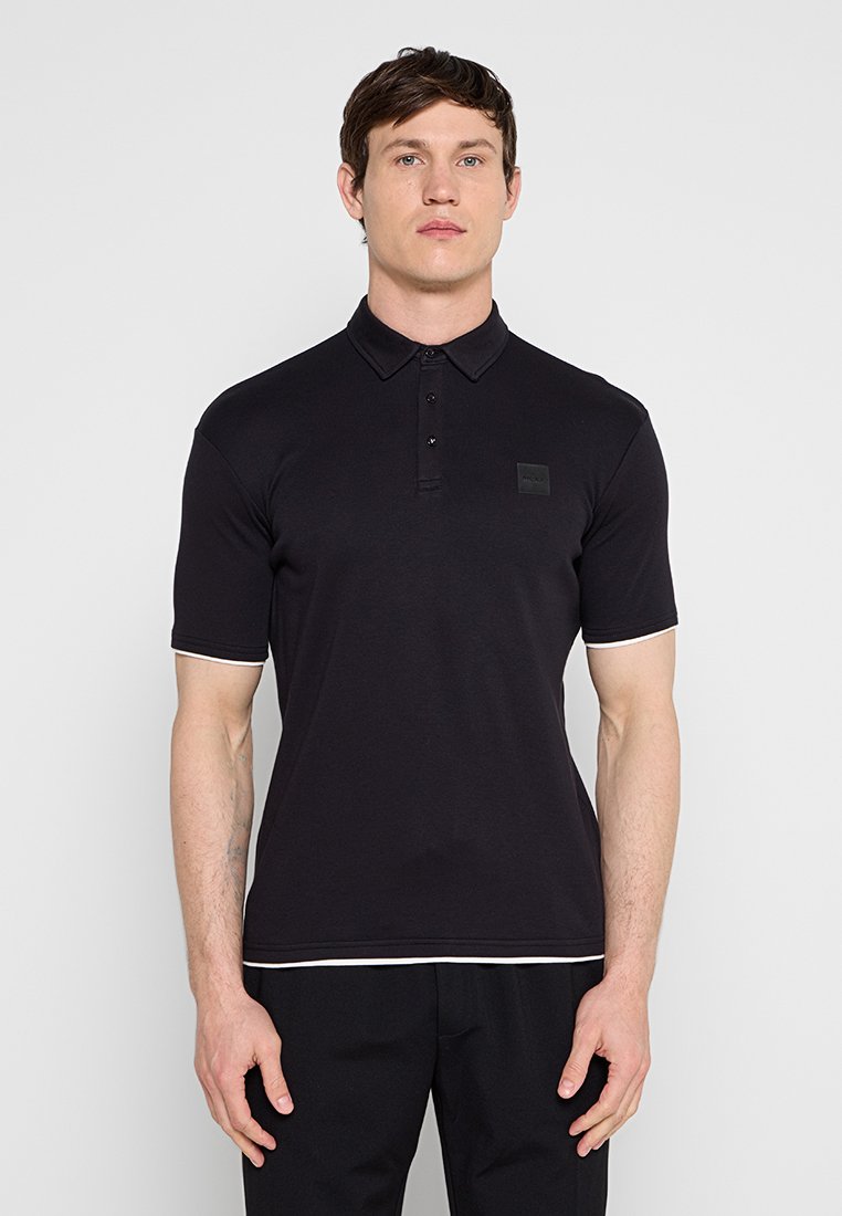 Mann steht und trägt ein schlichtes schwarzes Kurzarm-Poloshirt und schwarze Hose vor einem einfarbigen hellgrauen Hintergrund.