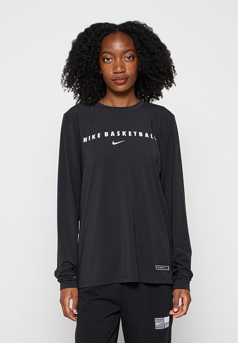 Frau mit lockigem Haar, die ein schwarzes langärmeliges Nike Basketball-Shirt und schwarze Hose trägt, vor einem einfarbigen Hintergrund stehend.